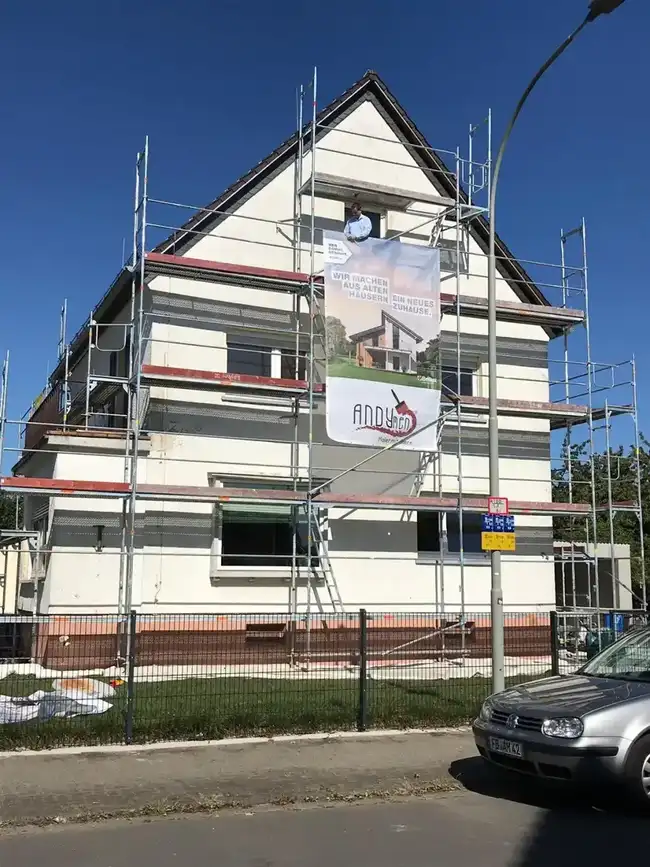 Fassade Einfamilienhaus Butzbach vorher — Geruest mit Andymen-Banner, Chef haengt Firmenschild auf